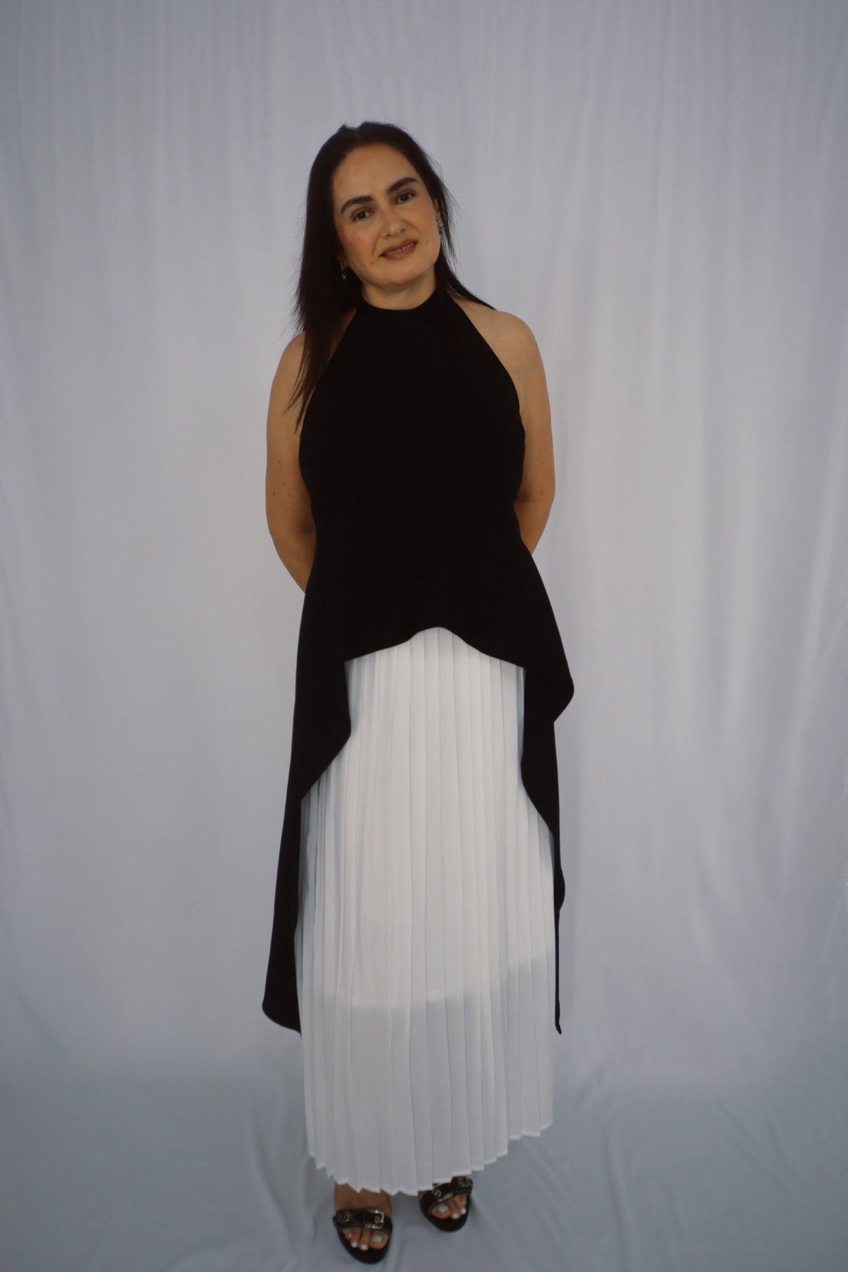 Model wearing a black asymmetric halter top with a high neckline and extended draped hem over a white pleated skirt.