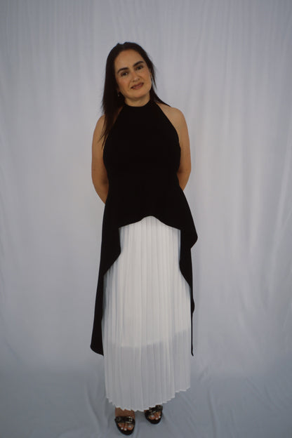 Model wearing a black asymmetric halter top with a high neckline and extended draped hem over a white pleated skirt.