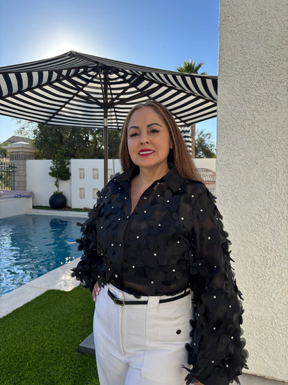 Woman wearing a black sheer blouse with 3D floral appliqué details and pearl accents, styled with white high-waisted pants outdoors by a pool.