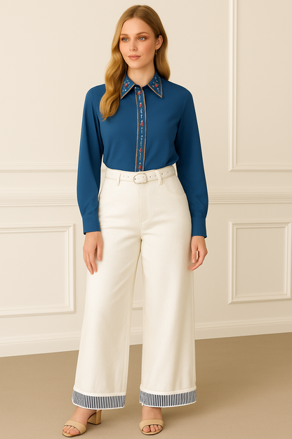 Woman wearing a blue shirt with a patterned collar and white pants against a beige wall.